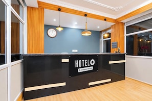 A hotel reception desk with a sign indicating it is company-serviced. There are two wall clocks, a window with a view, and pendant lighting. - Hotel O Chitrakut Nagar Royal Brothers (Danapur)