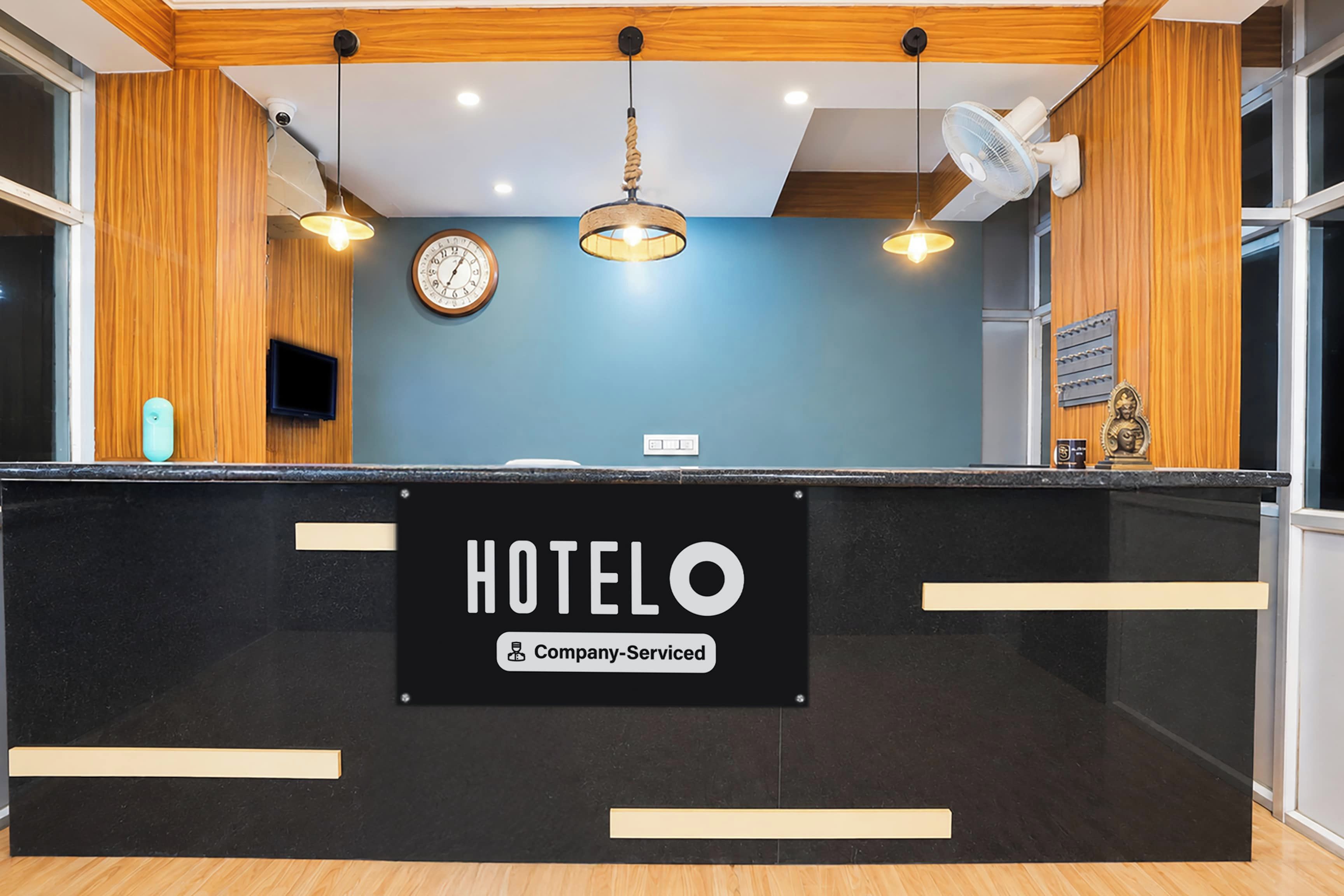 Bancada de recepção de hotel com relógio, placa indicando serviço da empresa e um ventilador fixado na parede.