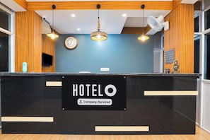 Bancada de recepção de hotel com relógio, placa indicando serviço da empresa e um ventilador fixado na parede.