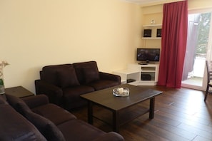 Ein Wohnzimmer mit einer Sofagarnitur, einem Couchtisch, einem Fernseher und einer Schiebetür zum Außenbereich.