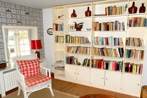 A room with a white wicker chair, a red and white checkered cushion, a white bookshelf filled with books, and a window with curtains. - Waldow im Ulmenhof 5 (Nieblum)