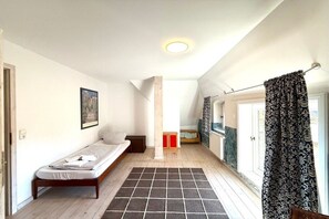 A small, single-bed room with a wooden floor, a black and white rug, a window with curtains, and a door leading to another room. - Holiday home in Oldsum with swimming pool (Oldsum)
