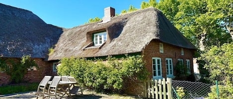 A thatched-roof house with a brick exterior, a wooden fence, and a garden area with chairs.