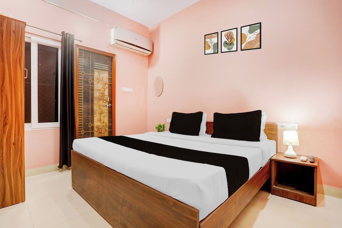 A bedroom with a bed, a nightstand, a lamp, a wall-mounted air conditioner, and a window with curtains.
