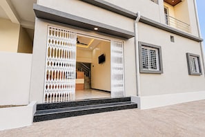 Edifício moderno com portão fechado, escadaria que leva à entrada e interior visível com uma televisão.