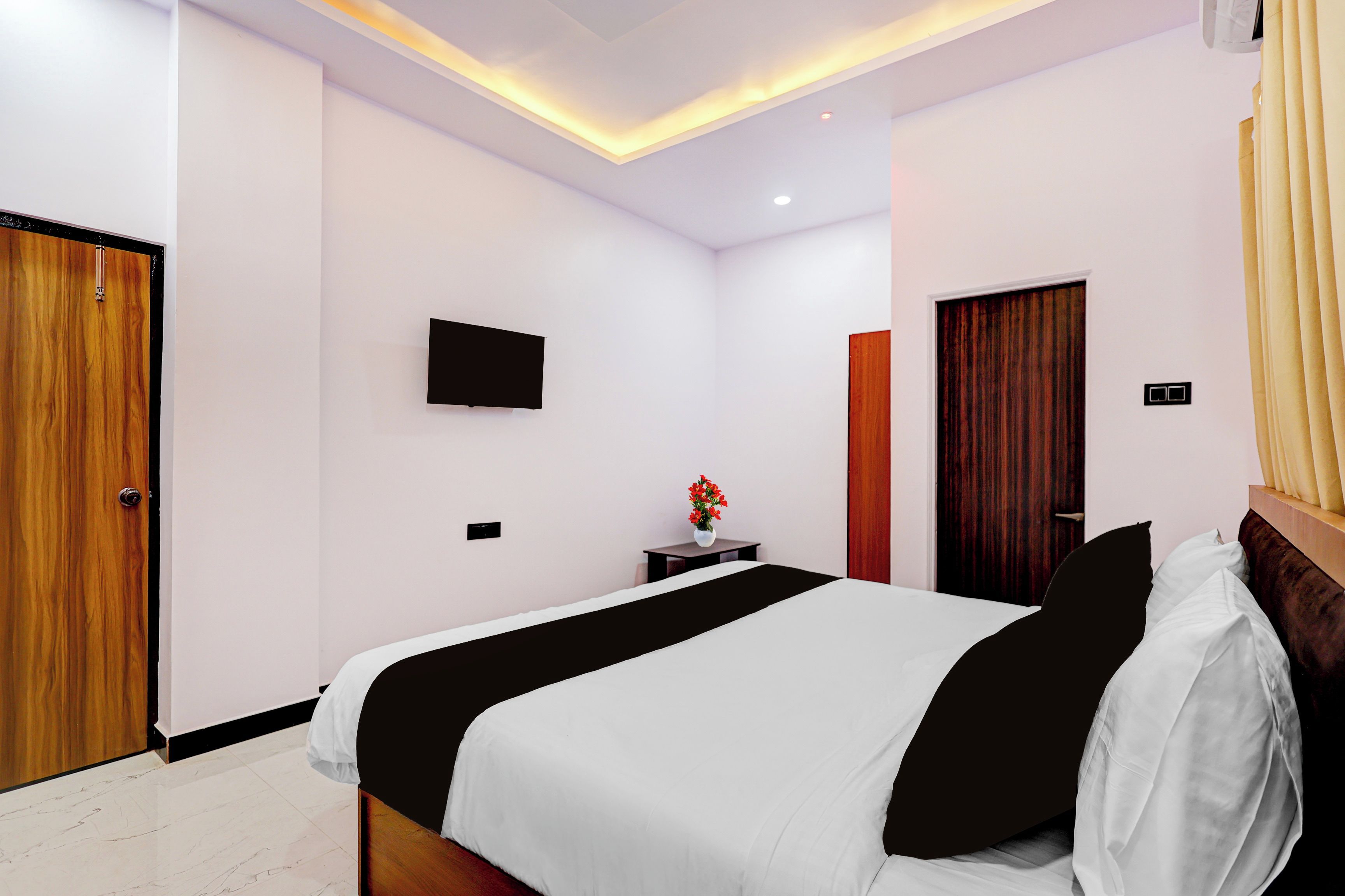 Una habitación de hotel con una cama, un televisor montado en la pared, una mesita con un florero y una puerta.