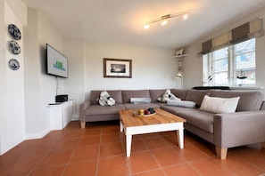 Ein Wohnzimmer mit grauem Sofa, einem Holztisch und einem an der Wand befestigten Fernseher.