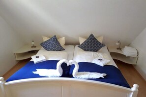 A double bed with a blue bedspread and white pillows, two bedside tables with lamps, and a heart-shaped towel decoration on the bed. - Holiday home at the spa park 1a (Wyk auf Föhr)