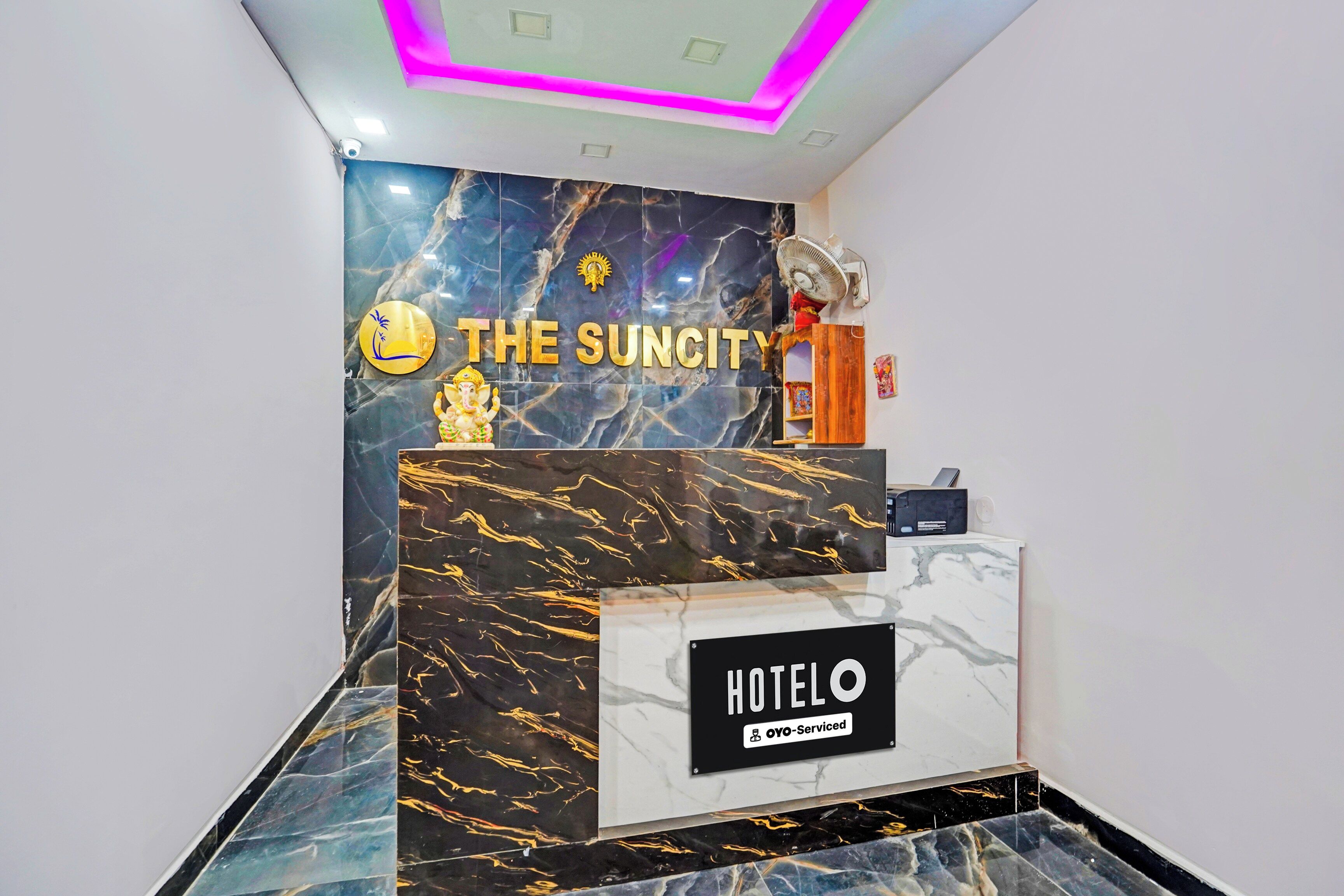 A reception desk with a marble wall, a sign reading 'THE SUNCITY HOTELO', and a fan on the wall.