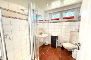 Et badeværelse med bruser, håndvask, toilet og et vindue med røde persienner.