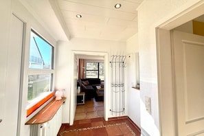 Ein Flur mit Heizkörper, Fenster mit Jalousien, einer Tür mit Metallgestell und Blick in einen Wohnbereich mit Sofa und Tisch.