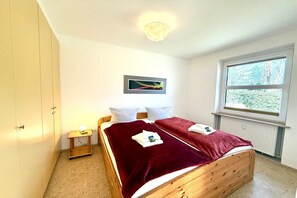 A bedroom with a wooden bed, a bedside table with a lamp, a window with a view of trees, and a framed picture on the wall. - Holiday apartment in Wyk on Föhr with sauna (Wyk auf Föhr)