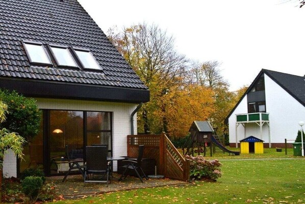 En boligejendom med skråt tag, en terrasse med udendørs møbler og et legeområde med rutsjebane.