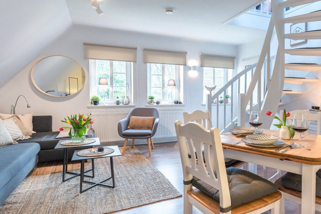 A modern living room with a sofa, a dining table set for two, a staircase, and a large mirror.
