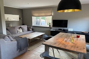 En moderne stue med en grå sofa, et træspisebord, et sort fjernsyn og et stort vindue med udsigt.