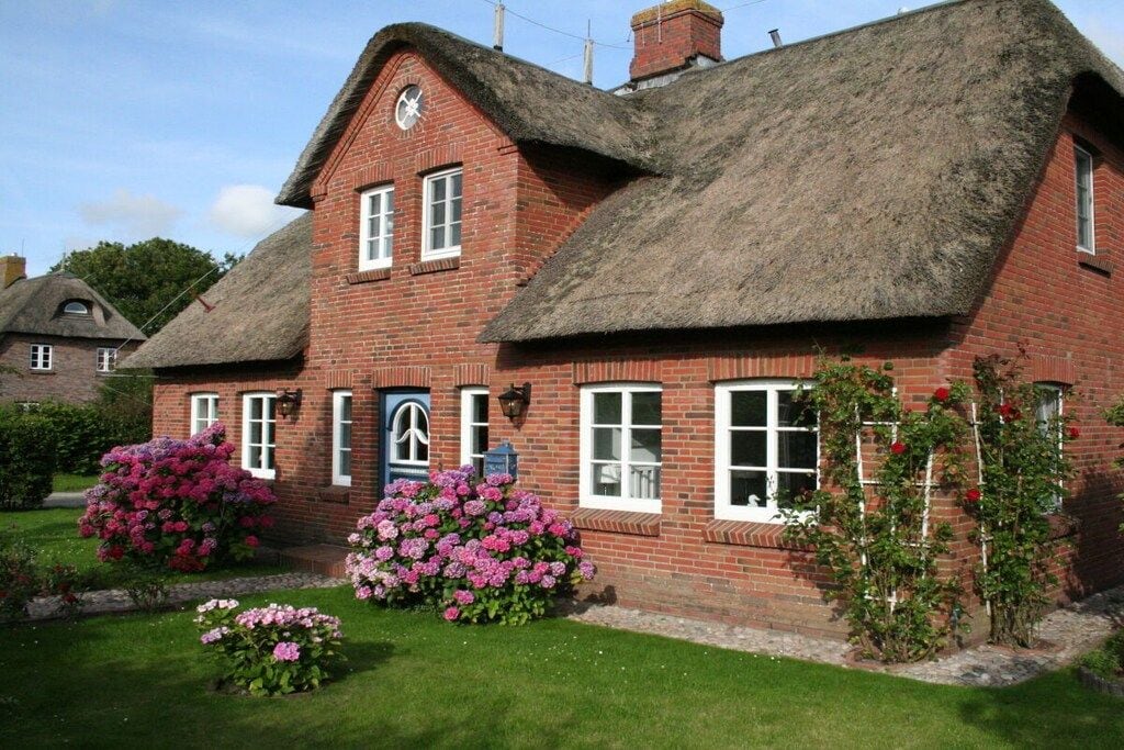 A traditional brick house with a thatched roof, a front door, and a small garden with pink flowers.