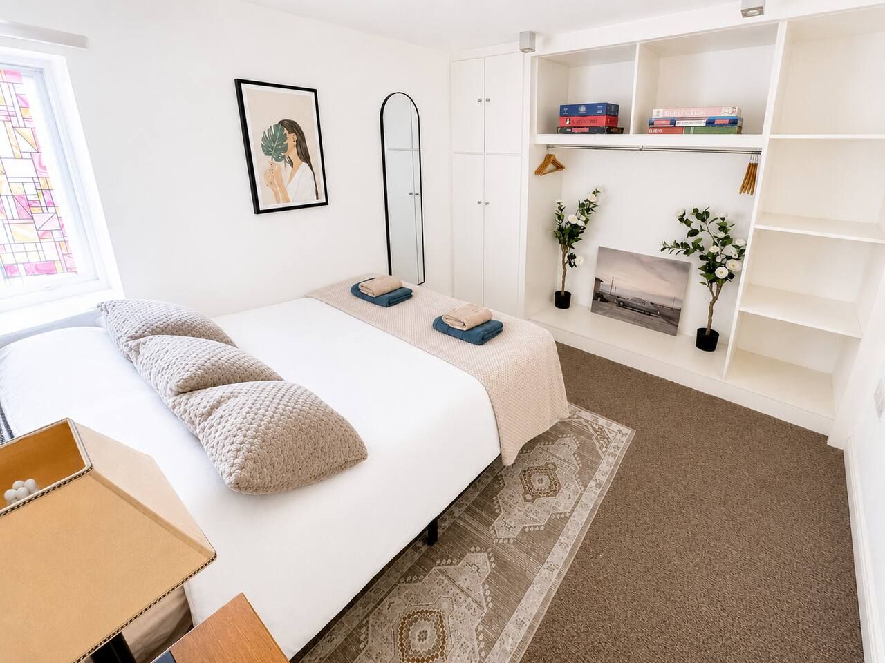 Een slaapkamer met een bed, nachtkastje, kledingkast, spiegel, boekenplank en raam.