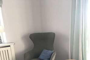 Ein Eckzimmer mit einem grau gepolsterten Sessel, einem blauen Kissen, einem weißen Heizkörper und einem fenster mit Vorhängen.