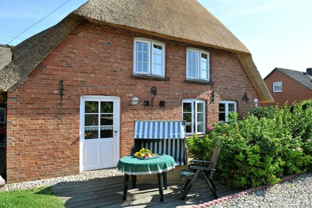 Une maison en briques avec un toit de chaume, une petite terrasse avec une table et des chaises, et un jardin.