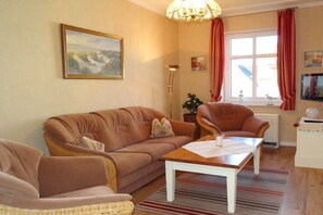 Una sala de estar con un sofá, un sillón y una mesa de centro. Hay un cuadro en la pared, una lámpara de pie y una ventana con cortinas.