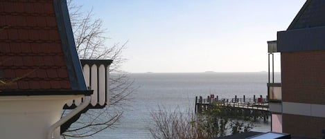 Vista costera con un muelle de madera que se adentra en el agua, cielo despejado y un edificio con techo inclinado.