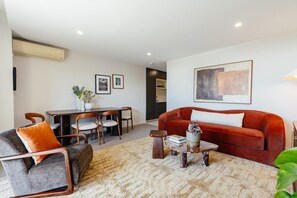 Een moderne woonkamer met een rode bank, een grijze fauteuil en een salontafel. Er is een eethoek met een houten tafel en stoelen.