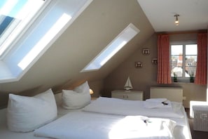 A room with a sloped ceiling, skylight, a bed with white bedding, a nightstand, a small sailboat decoration, and a window with curtains. - Haus Hansen Wyk - Holiday Apartment 03 (Wyk)
