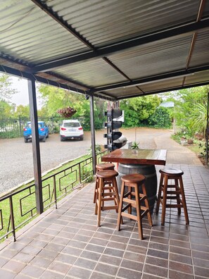 Une terrasse extérieure couverte, avec une table et des tabourets en bois, une rambarde en métal et une vue sur une route où circulent des voitures.