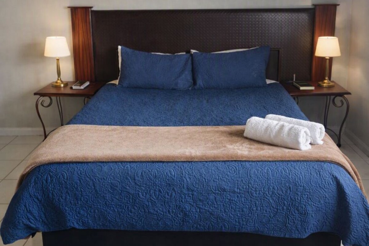 Un lit avec une tête de lit sombre, une literie bleue et un plaid marron. Il y a deux lampes de chevet et un tableau au-dessus du lit.