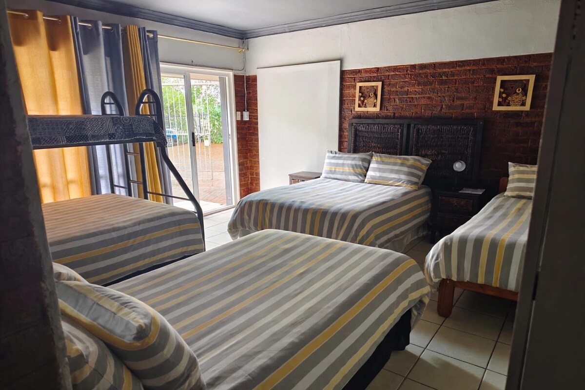 Une chambre d’hôtel avec trois lits simples, une porte coulissante donnant sur un balcon et un mur en briques orné de deux tableaux encadrés.