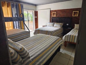 Une chambre d’hôtel avec trois lits simples, une porte coulissante donnant sur un balcon et un mur en briques orné de deux tableaux encadrés.