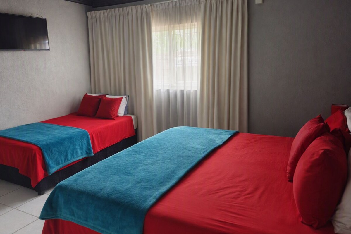 Une chambre d’hôtel avec deux lits, chacun étant équipé de draps rouges et d’une couverture bleue. Un téléviseur à écran plat est fixé au mur, et la chambre dispose d’une fenêtre avec des rideaux.