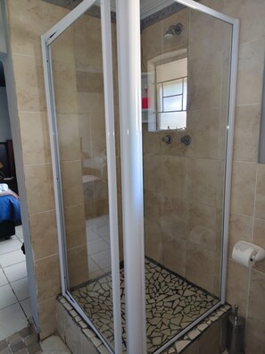 Une douche fermée par des parois de verre, avec un sol carrelé, un chambranle de porte blanc et une petite étagère sur laquelle se trouve un objet rouge.