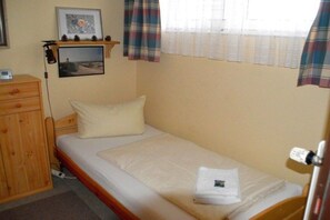 A single bed with a white mattress and pillow, a wooden nightstand with a lamp, a small shelf with decorative items, and a window with plaid curtains. - Holiday apartment in Wyk with swimming pool (Wyk)