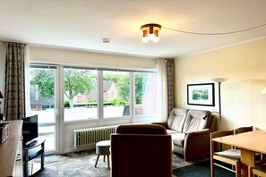 A hotel room with a sofa, a dining table, a TV, and a large window with a view of the outside. - Holiday apartment in Wyk with swimming pool (Wyk)