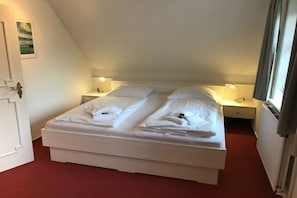 A double bed with white linens, two bedside tables with lamps, a door with a decorative handle, and a window with curtains. - Hansen Nieblum - Holiday Apartment 1 (Nieblum)