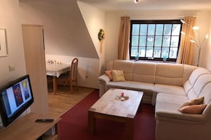 A living room with a sofa, a coffee table, a TV, and a window with curtains. - Hansen Nieblum - Holiday Apartment 1 (Nieblum)