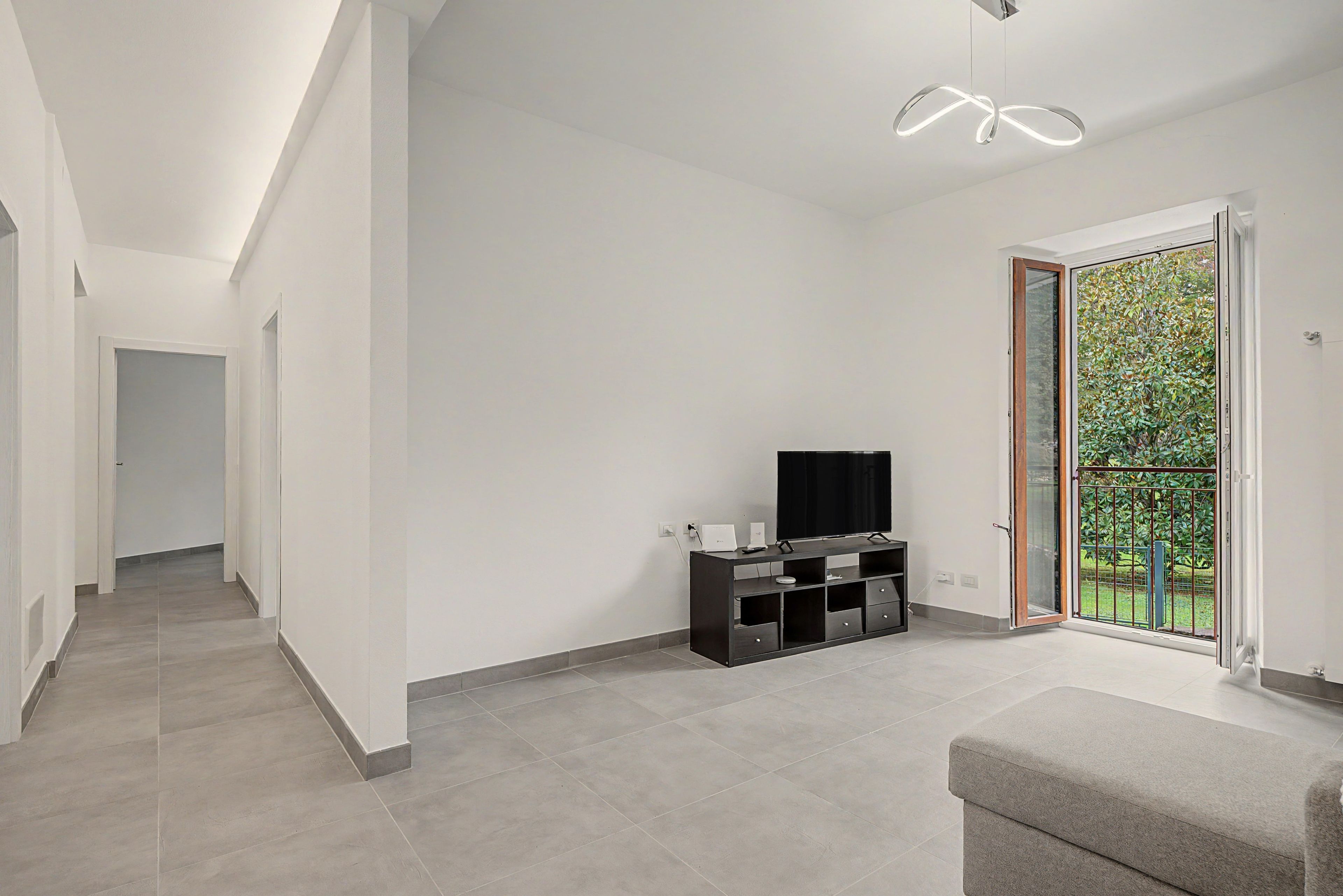 A modern living room with a flat-screen TV on a black entertainment unit, a grey sofa, and a sliding glass door leading to a balcony with greenery.