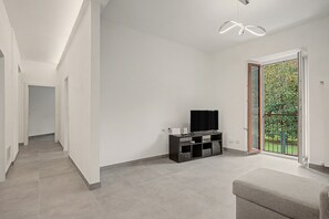 A modern living room with a flat-screen TV on a black entertainment unit, a grey sofa, and a sliding glass door leading to a balcony with greenery. - Holiday home 'Maison Du Lac' with balcony and Wi-Fi (Baveno)