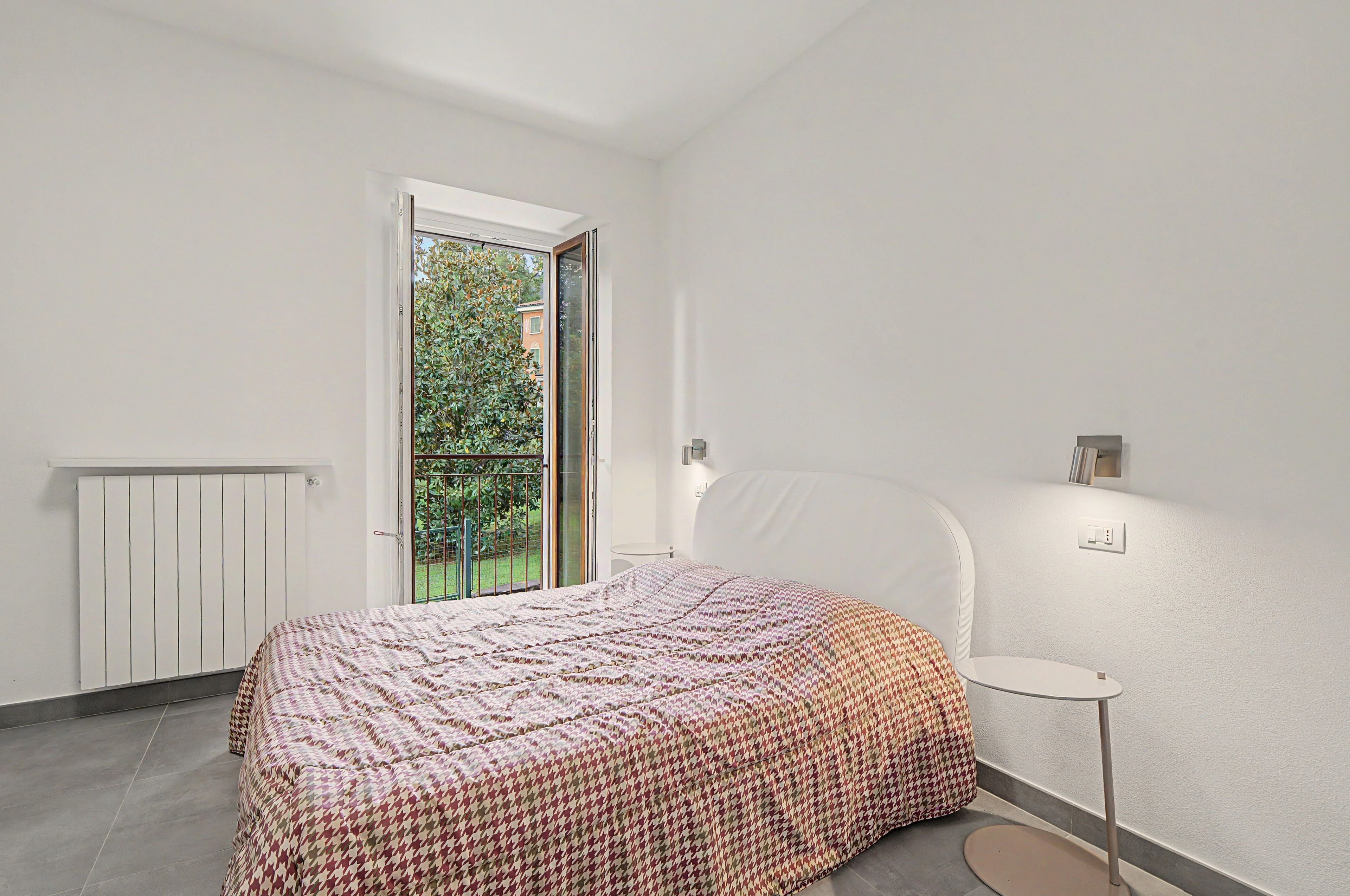 A bedroom with a bed, a small table, a radiator, and a window with a view of greenery.