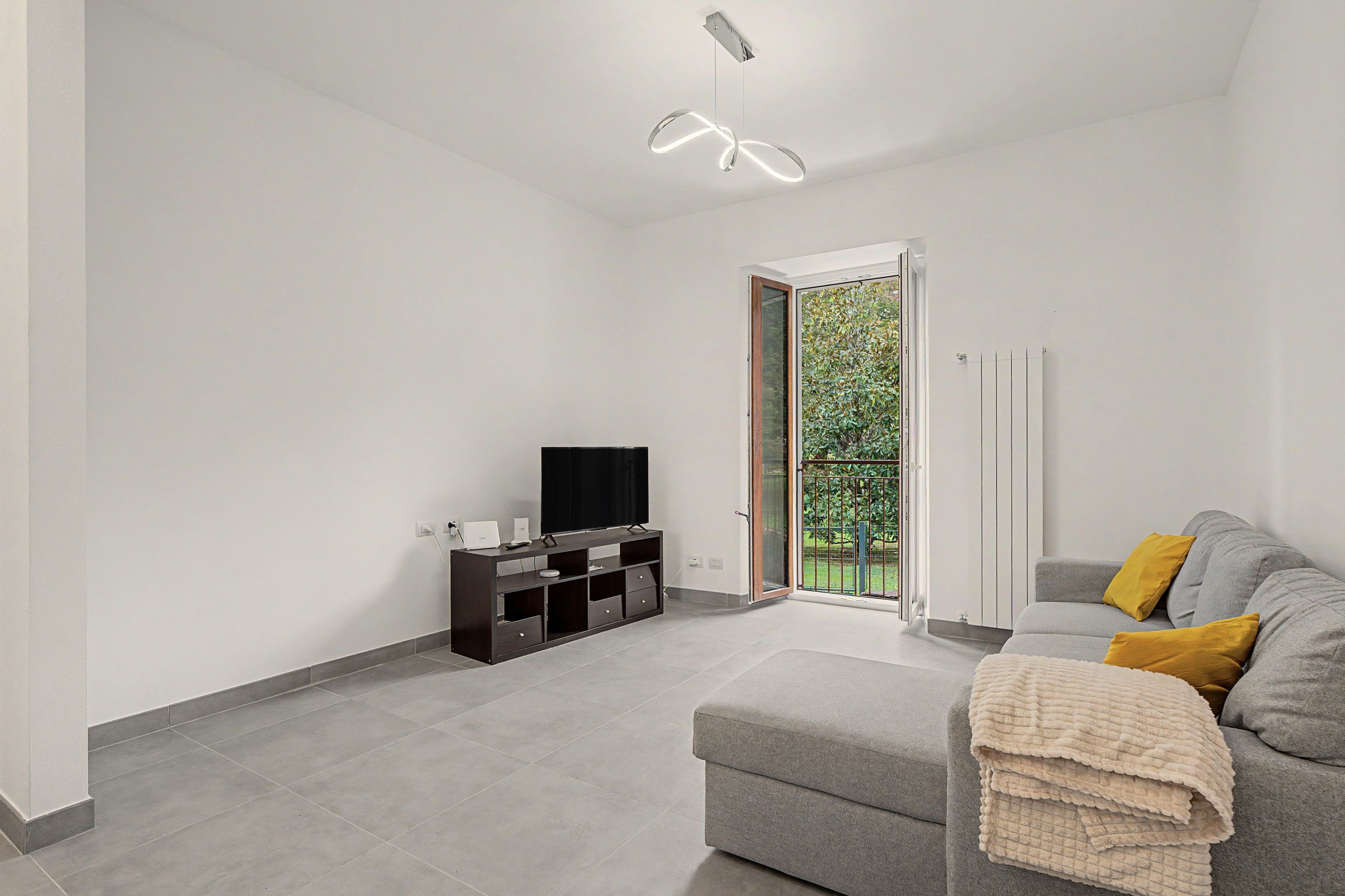 Ein modernes Wohnzimmer mit einem grauen Sofa, einem Flachbildfernseher auf einer dunklen Holz-Unterhaltungseinheit und einem Balkon mit einem Metallgitter.