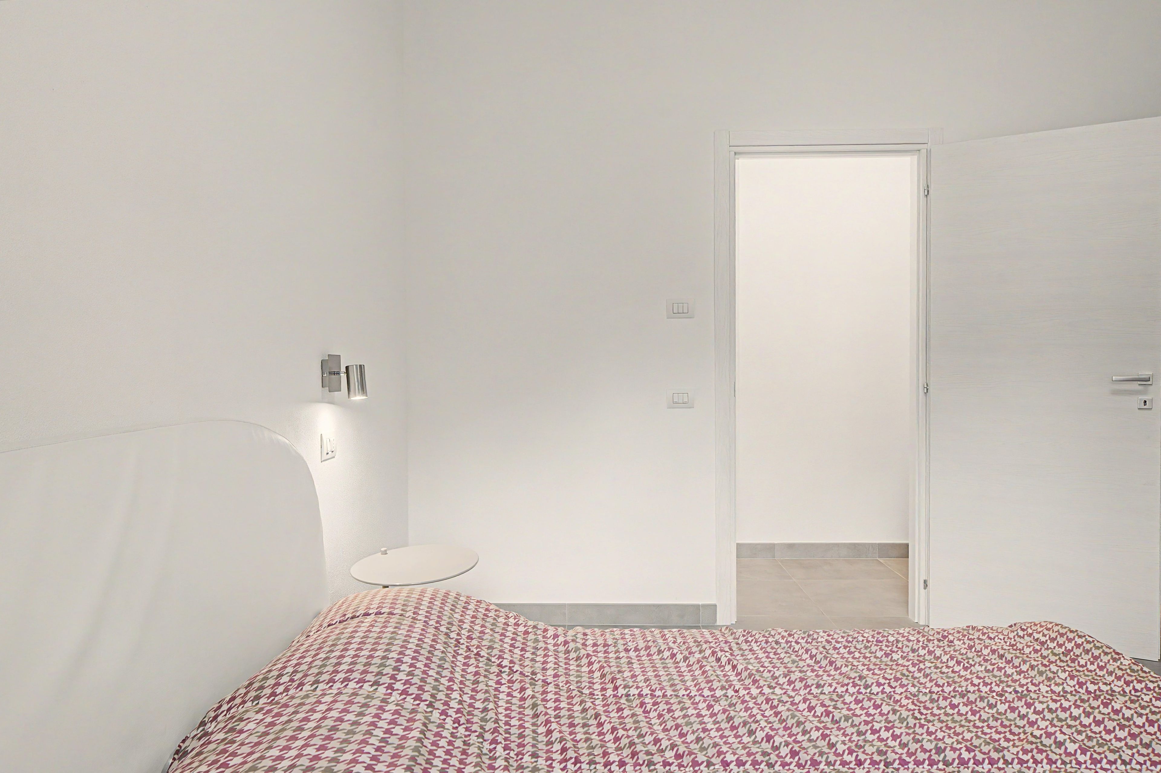 A minimalist bedroom with a bed, a bedside table, a wall-mounted light, and a door leading to another room.