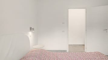 A minimalist bedroom with a bed, a bedside table, a wall-mounted light, and a door leading to another room.