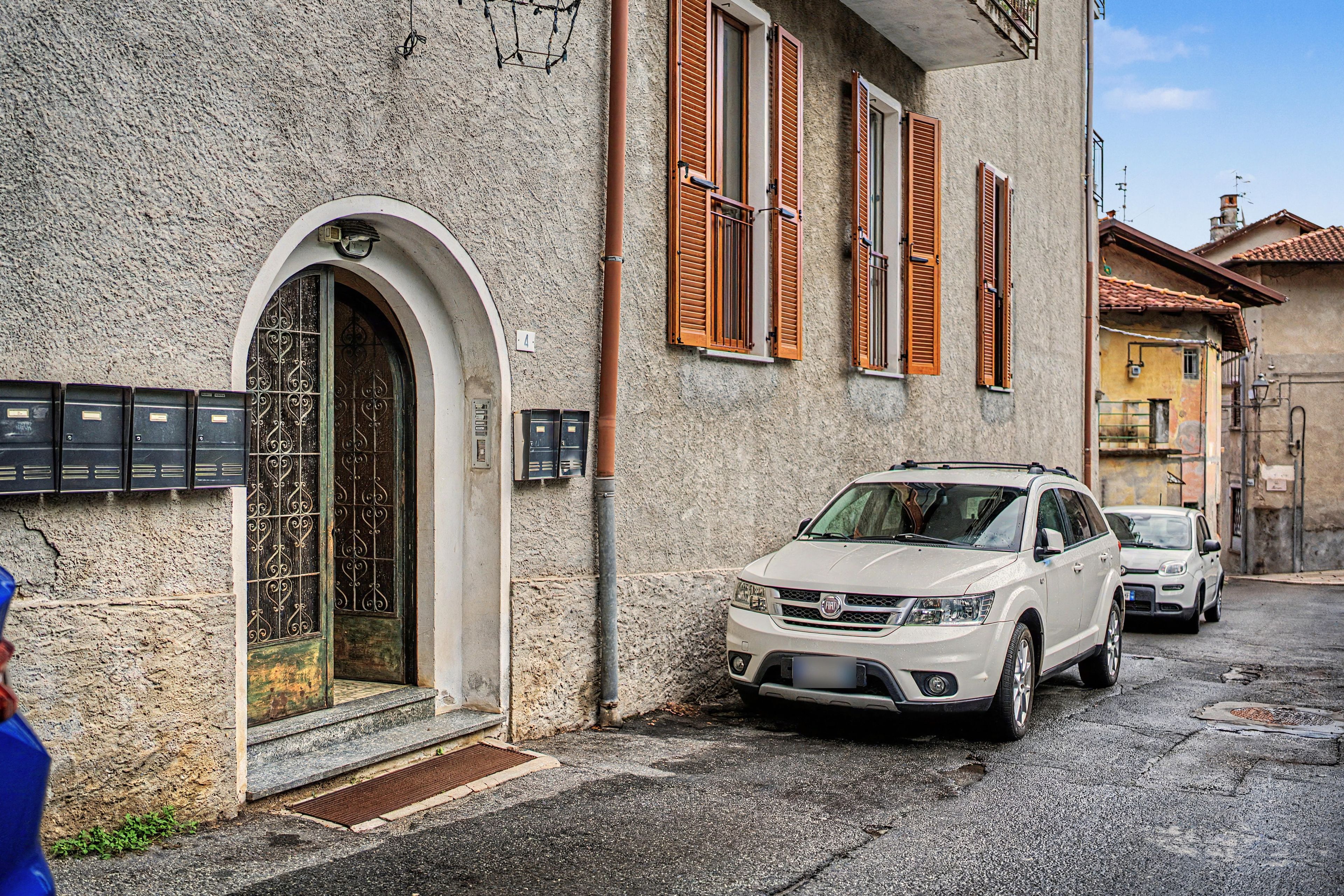 Un VUS blanc est garé devant un immeuble dont la façade est en pierre, avec des volets en bois et un portail en métal.