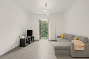 A modern living room with a grey sofa, a television on a black stand, and a large window with a view of greenery.