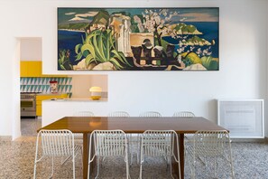 A dining area with a large painting, a kitchen in the background, and a table with chairs. - Exclusive villa with pool, 200 meters from the sea - DiCasaInSicilia (Syracuse)
