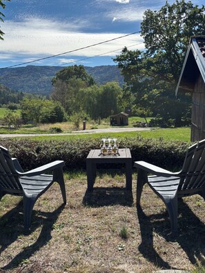 Two plastic chairs facing a table with a glass set, overlooking a rural landscape with a wooden structure and a hill in the distance.