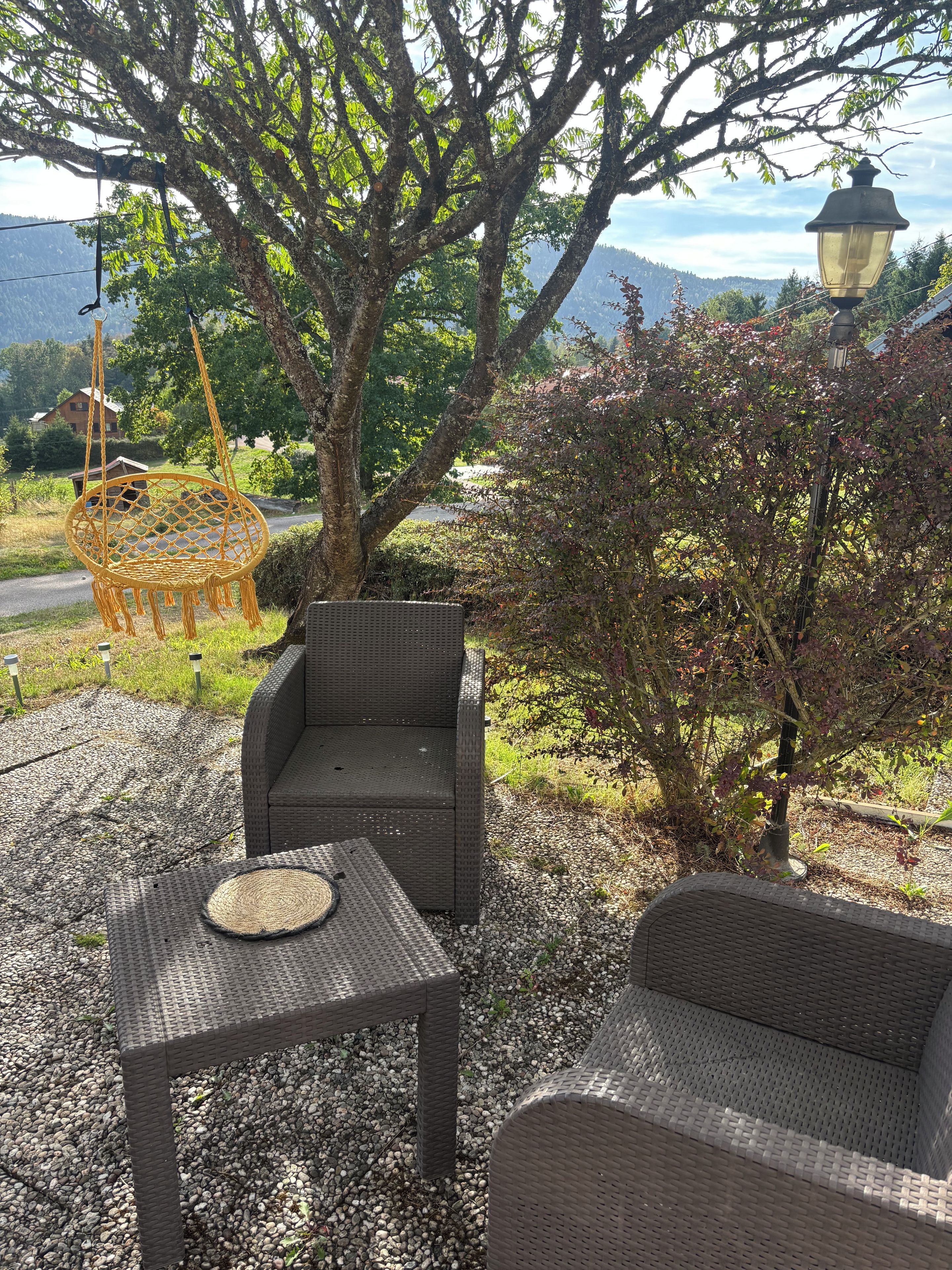 Ein Sitzbereich im Freien mit Korbmöbeln und einem Hängesessel, umgeben von Bäumen und Blick auf ferne Hügel.