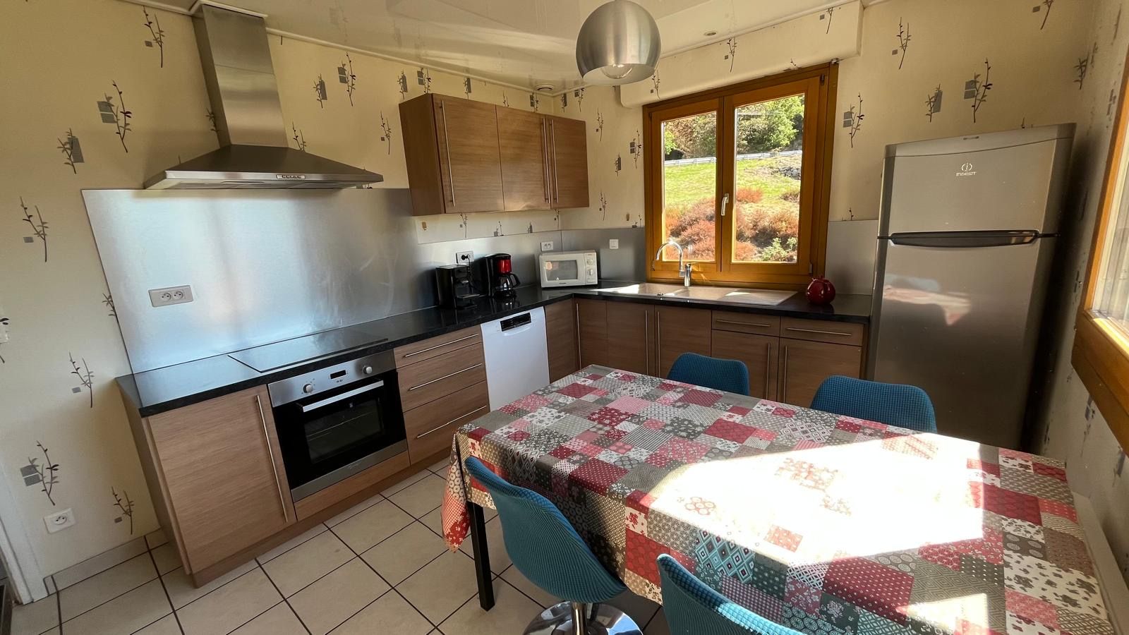 A compact kitchen with a stove, oven, microwave, and refrigerator. There is a dining table with chairs and a window with a view of greenery.