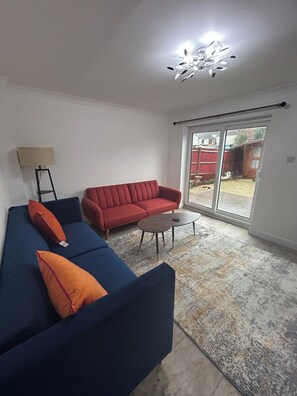 Een woonkamer met een blauwe bank, een rode bank, een klein rond tafeltje en een vloerlamp.